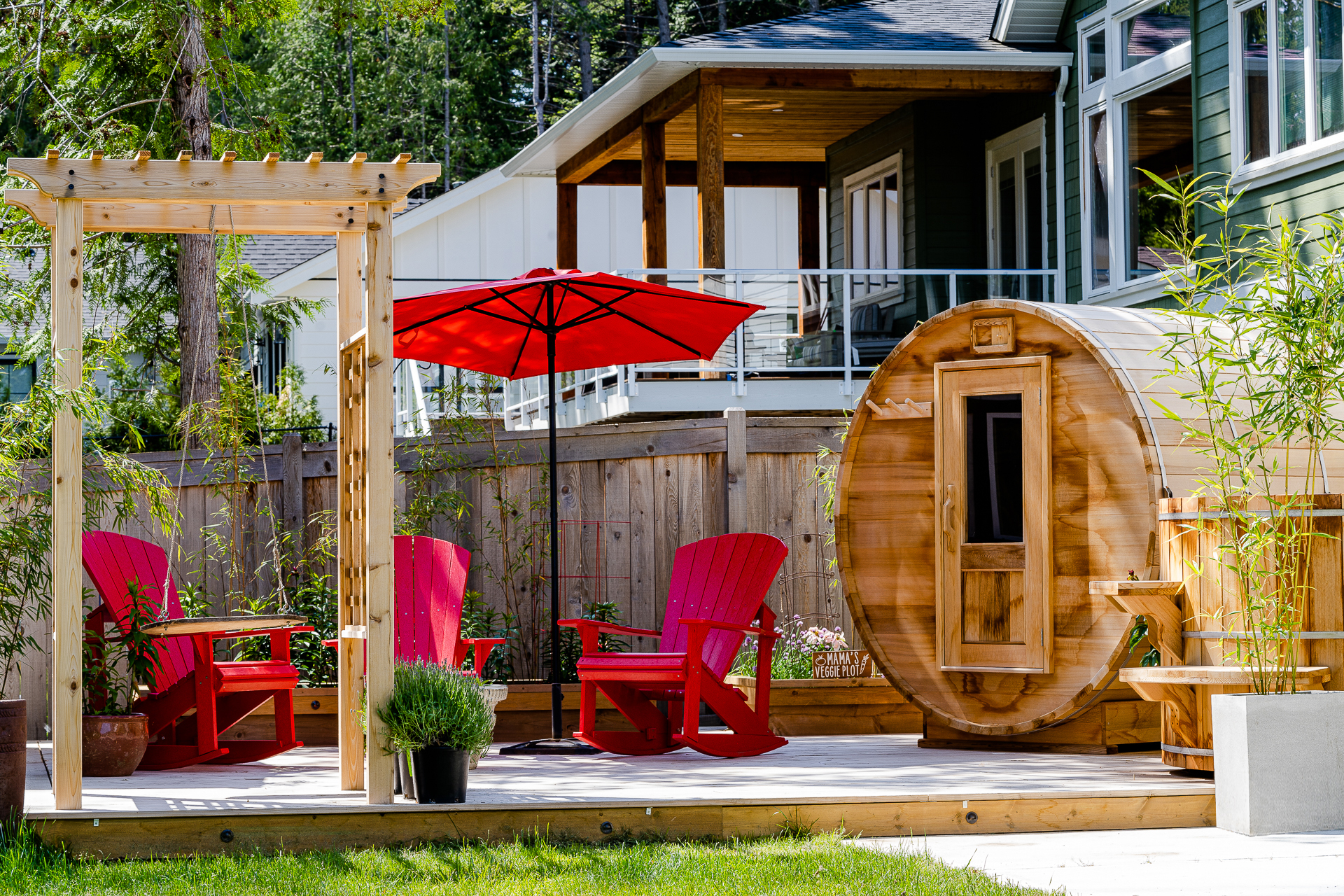 A beautifully designed backyard showcasing a cedar barrel sauna by Forest Cooperage. The scene includes a wooden pergola, vibrant red Adirondack chairs, and a red patio umbrella, creating an inviting outdoor space. This setup highlights the premium quality and aesthetic appeal of the cedar barrel sauna, ideal for relaxation and enhancing modern outdoor living.