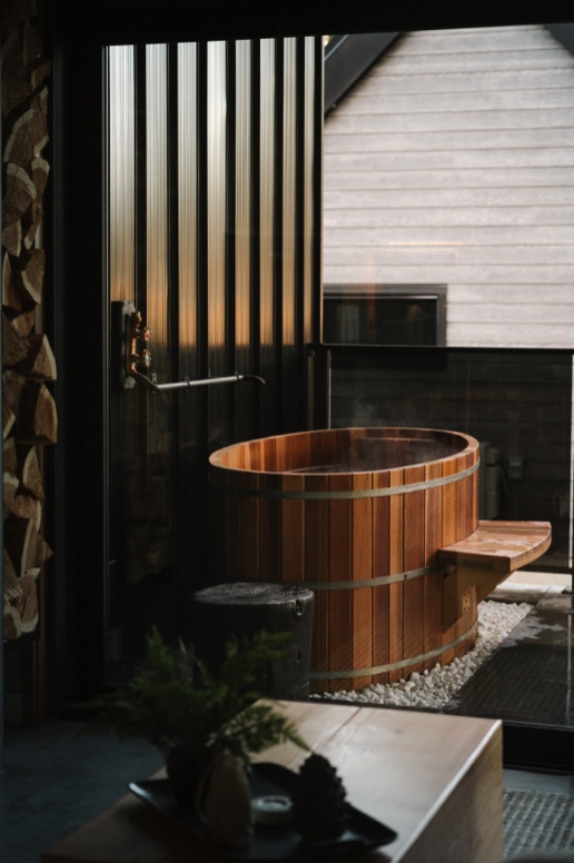 Japanese-style cedar soaking tub with traditional coopered design, located in a modern indoor-outdoor setting. The tub is nestled against a sleek black corrugated wall with warm natural light filtering through, highlighting the wooden craftsmanship